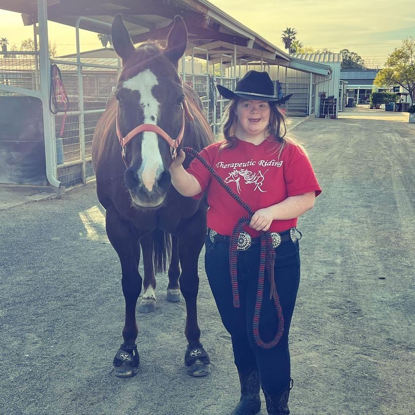 A rider in a Therapeutic Riding shirt leading her horse at sunset